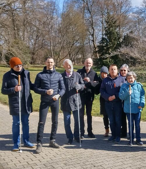 Menschen mit und ohne Sehbehinderung stehen in einem Park, drei von ihnen halten einen Blindenstock in der Hand.