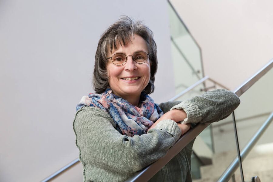 Monika Schaufler hat glattes, kinnlanges braungraues Haar. Sie trägt eine Brille mit schwarzer Umrandung und lächelt in die Kamera. Ihre Arme hat sie auf einem schrägen Treppengeländer abgelegt. Sie hat eine grünmelierte Jacke und einen bunten Schal an.