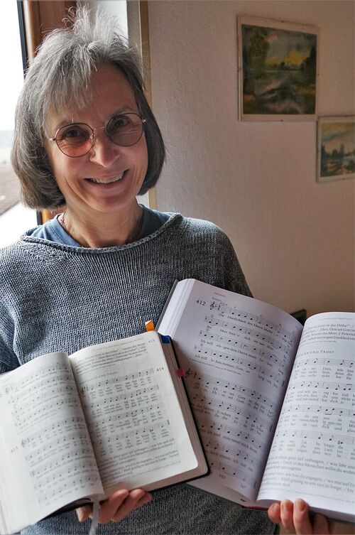 Monika Schaufler lächelt in die Kamera. Sie hat glattes, kinnlanges braungraues Haar und trägt eine Brille. In ihren Händen hält sie verschieden große, aufgeschlagene Ausgaben des Gotteslobes.