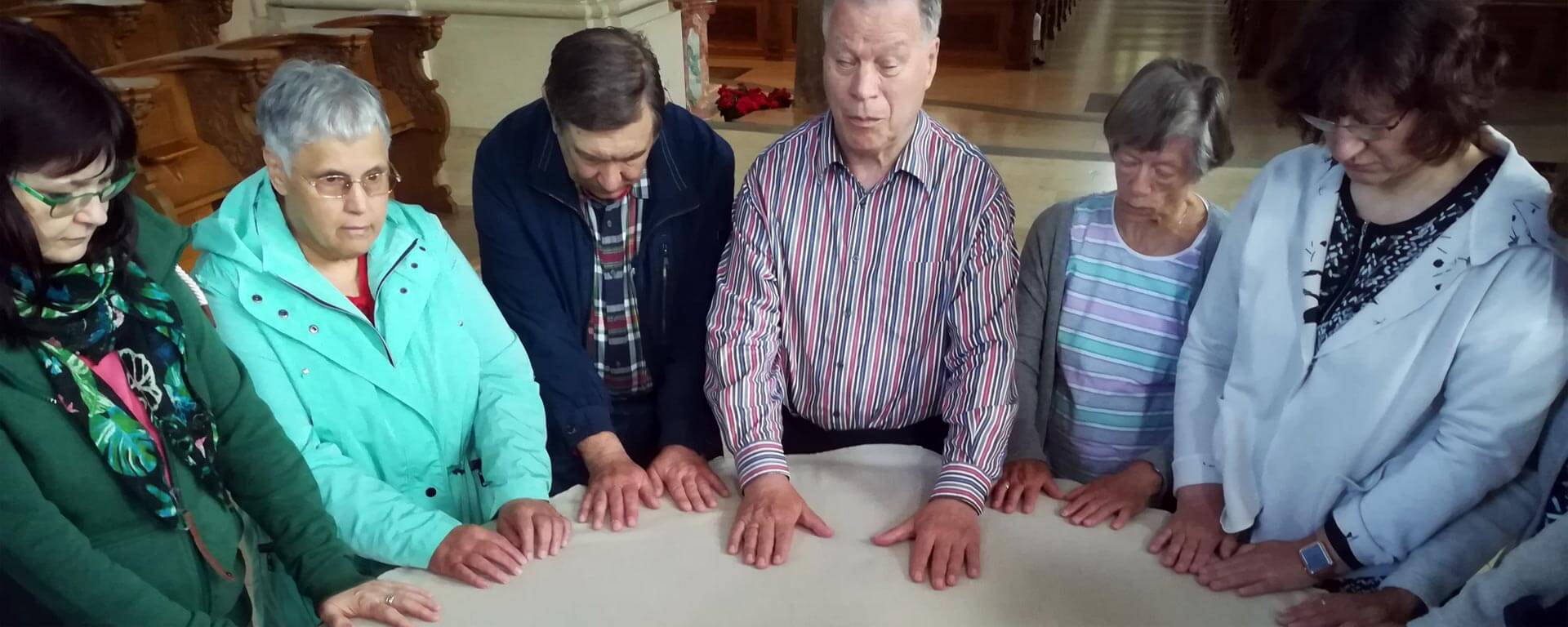 Menschen mit und ohne Sehbehinderungen legen ihre Hände auf den runden Altar in einer Kirche. Sie wirken dabei sehr konzentriert.