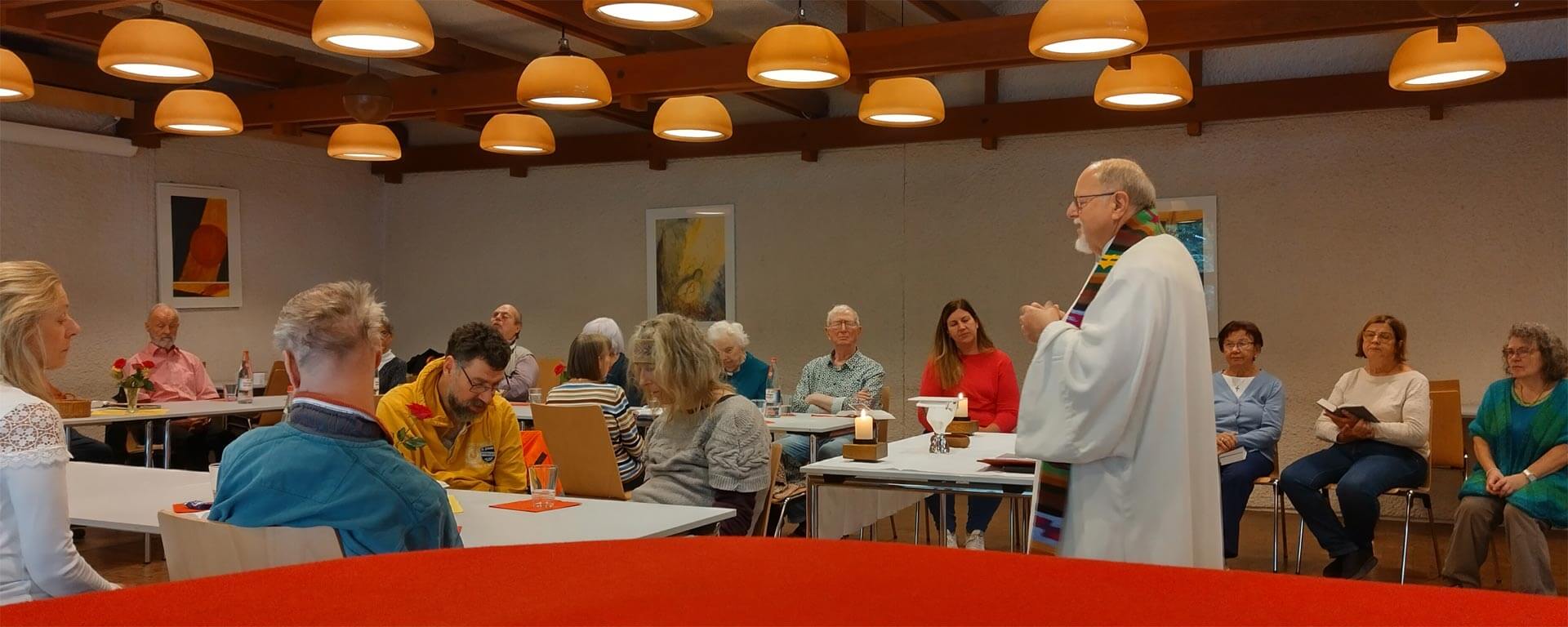 Eine Gruppe Menschen mit und ohne Sehbehinderungen sitzen in einem Halbkreis im Raum und lauschen der Ansprache des Pfarrers, der vor einem Tisch/Altar mit zwei brennenden Kerzen steht.